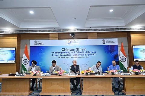 A panel of six individuals is seated at a long table adorned with flowers, flanked by Indian flags. A backdrop displays the event title about strengthening India's medical device export ecosystem, held in New Delhi on March 14, 2023. The setting suggests a formal, collaborative atmosphere.