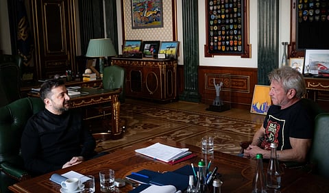Image of President Volodymyr Zelenskyy sitting across three time Oscar winner Sean Penn in Kyiv, Ukraine. The background is in a luxurious office with ornate wooden furniture and decorated walls. A table with documents and water bottles is between them.