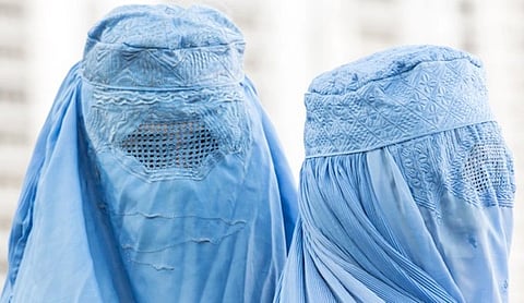 Two people in light blue burqas stand side by side. The garments feature intricate embroidery and mesh eye screens. The background is softly blurred.