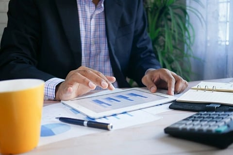 A person in a suit uses a tablet displaying bar graphs at a desk with a notebook, pen, calculator, and a yellow mug, conveying a business setting.