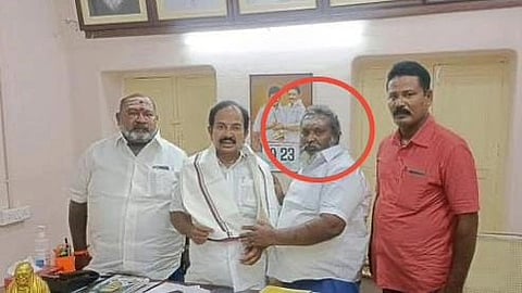 Four men from DMK pose in a room with framed portraits on the wall. One man is circled in red he is Senthur Karthu. The mood appears formal and serious. A table displays papers.
