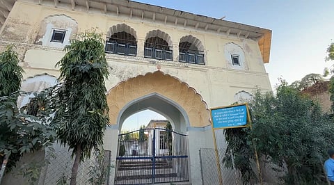 A quaint, colorful building with archways and intricate designs is framed by lush greenery. A small shrine stands at the entrance, conveying serenity.