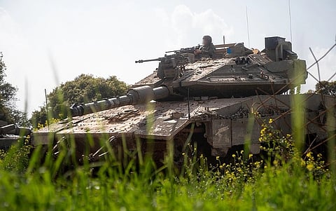 A tank partially hidden by tall grass in a sunny field. A soldier is visible on top of the tank, which is set against a backdrop of trees and blue sky.
