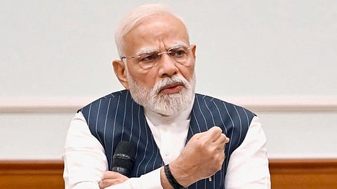 An image of PM Modi is shown wearing a striped navy vest over a white shirt, gestures emphatically while holding a microphone, conveying determination.