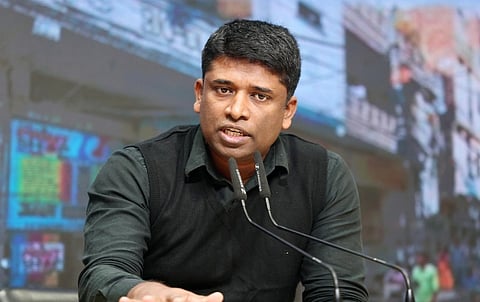 Image of former IAS officer Kannan Gopinathan speaking in front of a mic. the background of the image depicts a building and streets on a screen which is blurred.
