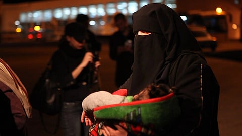 A woman in a black niqab holds a baby wrapped in a colorful blanket, standing in an outdoor setting at night. The scene conveys a caring and protective ambiance.