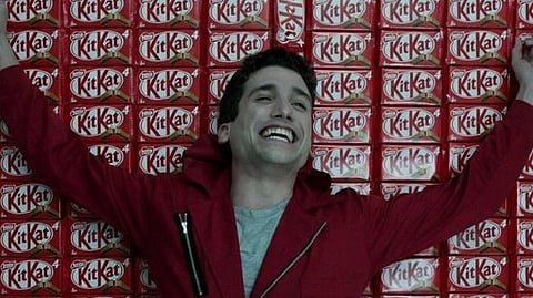 Smiling person in a red jumpsuit stands joyfully against a wall of stacked Kit Kat bars, conveying a playful and spirited mood.