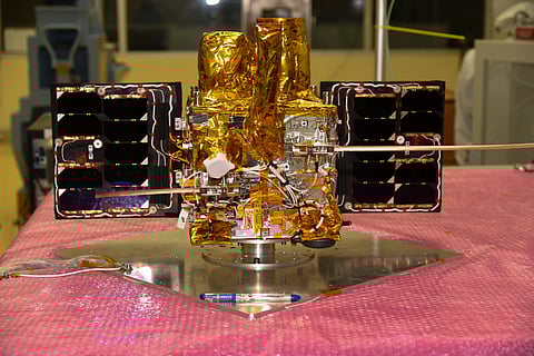 An experimental nano satellite developed by ISRO. gold foil covered satellite with side panels on a stand.