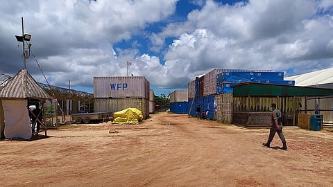 A dusty yard with blue and white containers, marked "WFP," under a partly cloudy sky. A person in a mask walks on the right. Rugged and functional atmosphere.