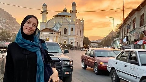 Shelly Kittleson in a black outfit and blue scarf stands on a busy street at sunset, smiling. Behind her is a grand mosque with a striking dome and minarets.