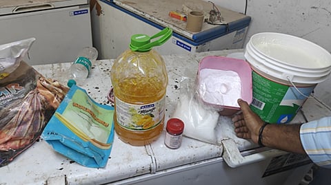 A cluttered countertop with a large oil bottle, spice packets, a water bottle, and an open tub of powder. A hand holds a pink tray over the tub.