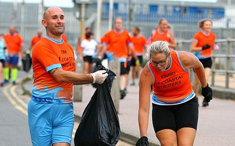 Two runners in orange "Coastal Striders" shirts collect trash in bags during an outdoor event, smiling and active. Others in matching shirts run behind them.