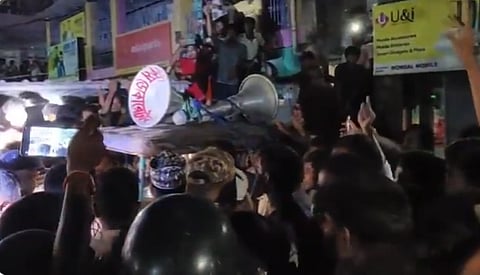 A bustling nighttime street scene with a dense crowd. People raise phones and hands toward a large speaker held aloft, capturing the lively, festive atmosphere.