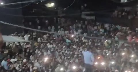 A large, lively crowd gathers outdoors at night, many holding phone lights. A person on stage, seen from behind, addresses the audience, creating an energetic atmosphere.