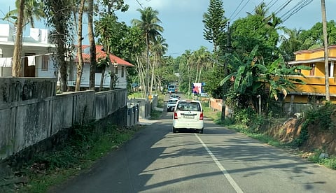 A narrow road flanked by houses and lush greenery, with palm trees lining the sides. Cars travel down the road, conveying calm and tropical ambiance.