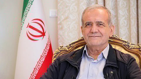 In the image Masoud Pezeshkian is shown sitting in a chair behind him is the Iran flag