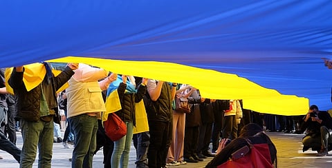 People stand under a large Ukrainian flag, the blue and yellow colors dominating the image, conveying unity and patriotic solidarity.