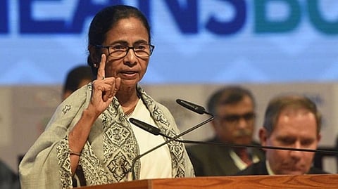 West Bengal CM, Mamata Banerjee in a patterned shawl and glasses speaks at a podium with a serious expression, gesturing with her hand.