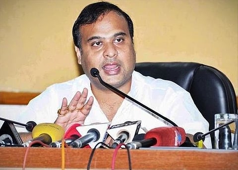 Himanta biswa sarma speaking at a press conference with multiple microphones in front. He is gesturing with one hand, conveying a serious and focused tone.