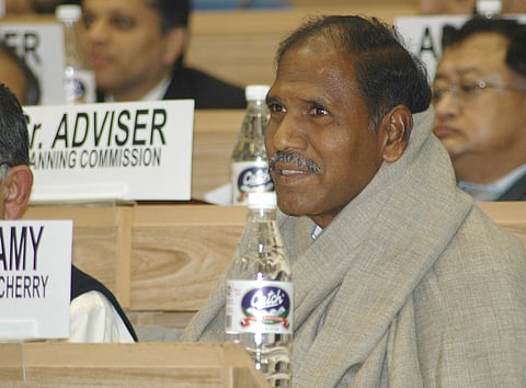 N Rangasamy sitting on a bench in a shawl, surrounded by nameplates and people, with a water bottle in front of him