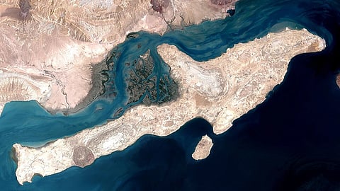 Satellite image of the Strait of hormuz, an arid coastal region with rugged, beige terrain. The land is surrounded by dark blue water, and there are intricate patterns of inlets.
