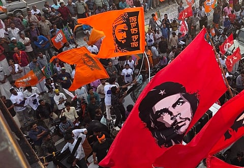 Aerial view of a large crowd of people holding red and orange flags with revolutionary symbols and figures of Chhatrapati Shivaji and Che Guevara. The atmosphere is lively and dynamic.