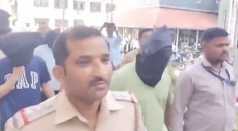 Police are escorting two individuals with covered heads outdoors. Officers in uniform lead and follow, creating a tense, secure atmosphere.