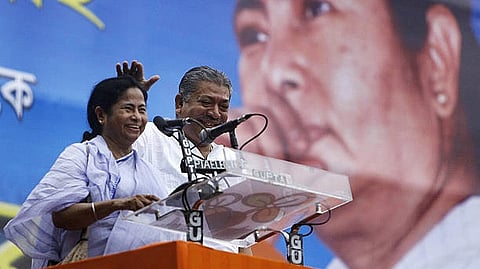 mamata banerjee stands in front of a poster with her face, behind a podium, next to a man