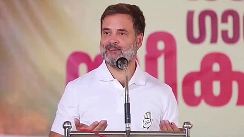 Rahul Gandhi in a white shirt, speaks at a podium with a microphone. Behind him, a blurred, colorful banner. He appears confident and engaged.