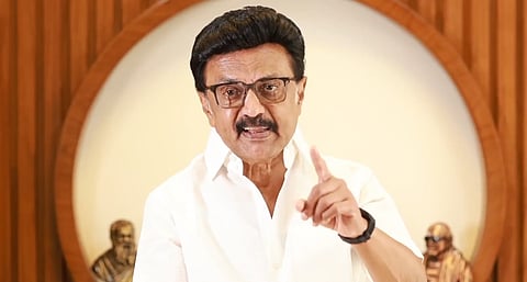 Image of MK Stalin in a white shirt speaks emphatically, pointing for emphasis. Behind him are wooden sculptures.