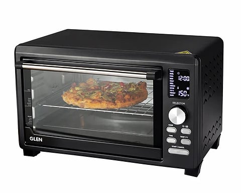 Black countertop oven with digital display showing 12:00 and 150°C. A pizza with toppings bakes on a wire rack inside, creating a homely atmosphere.
