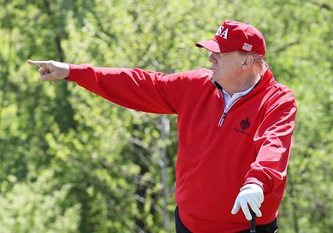 US President Donald Trump in a red "USA" cap and jacket points across a green, wooded golf course. They appear focused and engaged, with a golf glove on one hand.