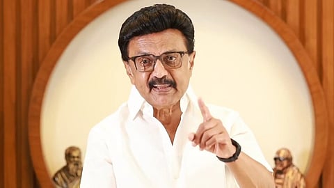 Image of MK Stalin in a white shirt speaks emphatically, pointing for emphasis. Behind him are wooden sculptures.