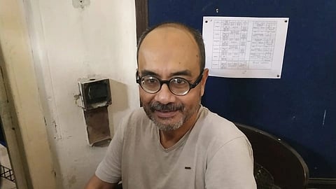Ashoke Sen sits smiling at a desk cluttered with papers. Behind him, a blue bulletin board with a pinned paper is visible.