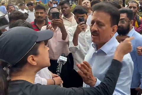 A woman wearing a cap is appearing to argue with a man in a blue shirt. A crowd of people surrounds them, many holding up phones to record the interaction. The scene conveys tension and public engagement.