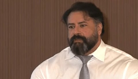 A man with a beard and mustache wears a white shirt and gray tie, seated in front of a brown backdrop. He appears thoughtful, with a microphone visible.