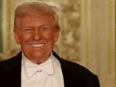 Image of Trump smiling warmly in a formal setting, wearing a tuxedo with a white bow tie. The background is elegant, featuring ornate, light-colored decor.