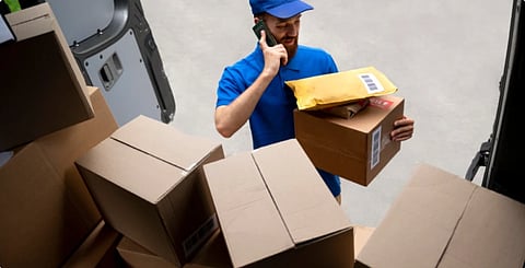 A delivery worker in a blue uniform holds a phone and a package inside a van full of boxes, conveying a busy and focused atmosphere.