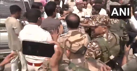 A crowded street scene shows military personnel in camouflage amidst a group of civilians near a vehicle. People appear tense and engaged, conveying urgency.