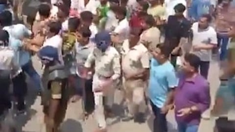 A large crowd of people, including police officers and civilians, walk together on a street in daylight. Some officers wear uniforms, creating an active and tense atmosphere.