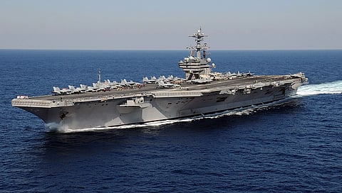 A large aircraft carrier sails on a calm blue ocean against a clear sky. Jets are lined up on deck, indicating military readiness and strength.
