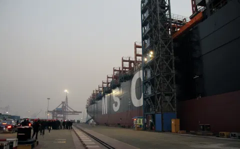 Cargo ship docked at a port during dusk, with cranes and shipping containers visible. People and vehicles are near the large ship, creating a bustling scene.