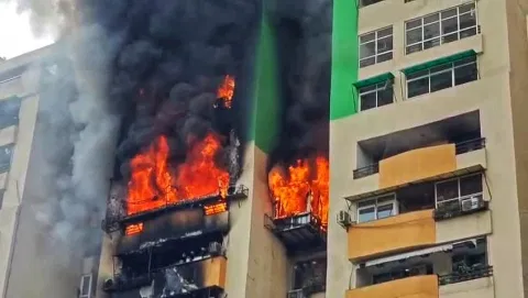 A high-rise building engulfed in flames, with black smoke billowing from multiple floors. The scene conveys urgency and danger.