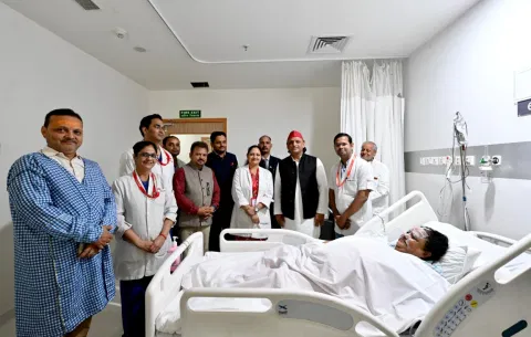 A group of people, including medical staff and visitors, stand around a patient lying in a hospital bed. The mood is supportive and attentive.