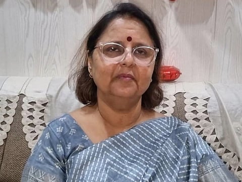 A woman with glasses and a bindi sits on a sofa, wearing a gray patterned saree. The background features a light-colored wooden wall and cushions.