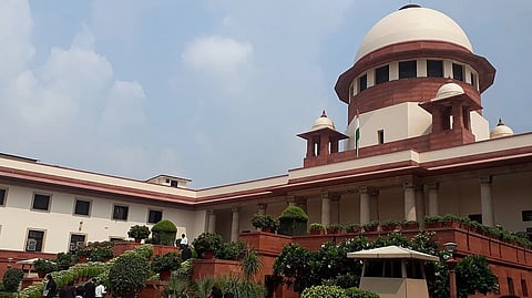 The image shows the Supreme Court of India's grand building under a blue sky. The architecture features a large dome, columns, and lush greenery, conveying authority.