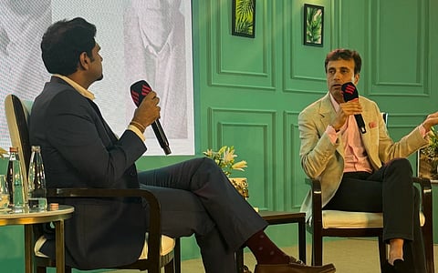 Two men in a discussion on stage, each holding a microphone. One wears a dark suit, the other a beige jacket. Background: green wall with art.