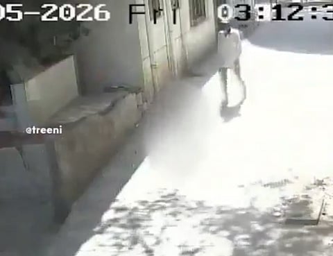A grainy CCTV image shows a person in light clothing walking on a sunlit street beside a building. The timestamp reads "2026," adding an unclear futuristic context.