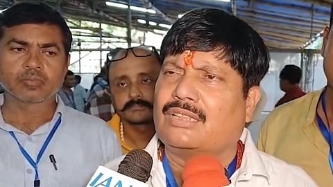 A man with a red tilak on his forehead speaks into microphones labeled "IANS" and another one. Two men stand blurred in the background. Indoor setting.