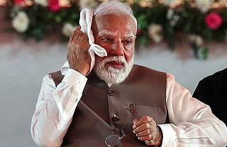 PM Modi in a brown vest wipes his brow with a white cloth, holding glasses, with a blurred floral background. He appears contemplative and slightly weary.